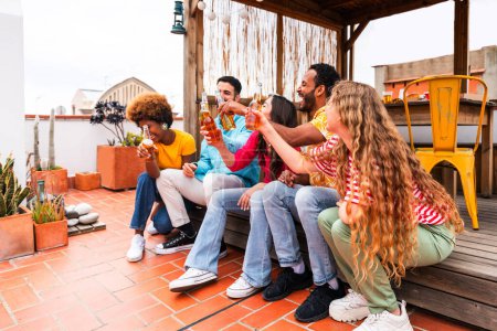 Photo for Multiethnic group of happy young friends having dinner barbecue party on rooftop at home - Multiracial cheerful young adult people having fun and bonding on a terrace balcony with city view, eating and drinking. - Royalty Free Image