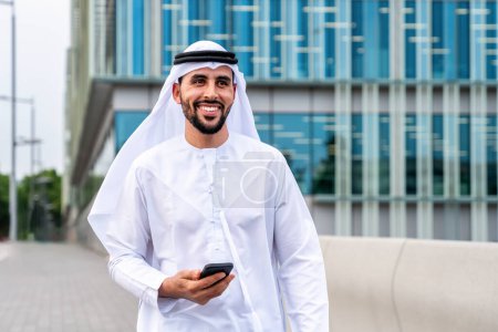 Foto de Hombre árabe de Oriente Medio vestido con ropa tradicional emirati kandora en la ciudad - Hombre de negocios árabe musulmán paseando por el centro de negocios urbano. - Imagen libre de derechos