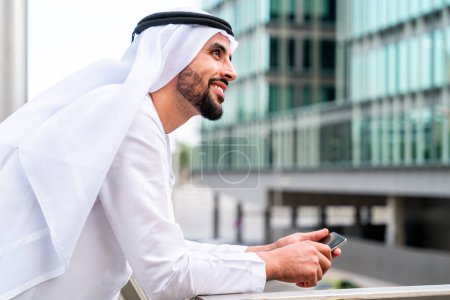 Foto de Hombre árabe de Oriente Medio vestido con ropa tradicional emirati kandora en la ciudad - Hombre de negocios árabe musulmán paseando por el centro de negocios urbano. - Imagen libre de derechos