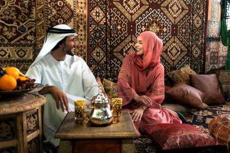 Photo for Young couple from Emirati spending time in an arabian traditional cafe. Man and woman wearing kandura and abaya from Dubai conversating together. - Royalty Free Image