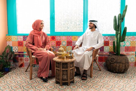 Photo for Young couple from Emirati spending time in an arabian traditional cafe. Man and woman wearing kandura and abaya from Dubai conversating together. - Royalty Free Image