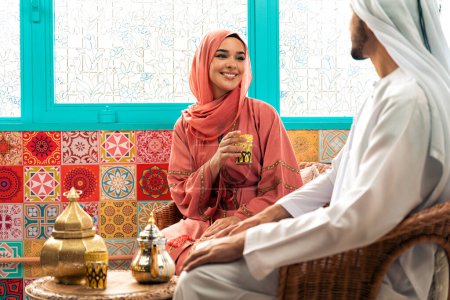 Téléchargez les photos : Un jeune couple émirien passe du temps dans un café arabe traditionnel. Homme et femme portant kandura et abaya de Dubaï conversant ensemble. - en image libre de droit