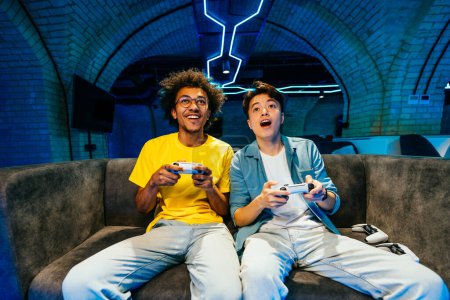 Foto de Grupo multiétnico de jóvenes amigos jugando videojuegos en casa sentados en el sofá - Videojuegos divertidos jugando deportes electrónicos en la consola de videojuegos - Imagen libre de derechos