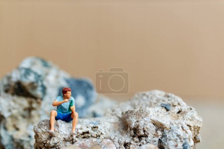 Miniaturmenschen, ein junger Mann nippt am Bier und raucht Zigaretten, während er auf dem Felsen sitzt