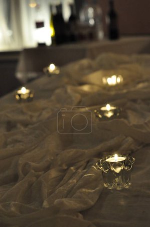 Photo for Arrangement of candles on a table surrounded by white fabric. Tealight candles in clear glass cups in dim lighting and romantic, ambient feeling. Small flames burn brightly. - Royalty Free Image