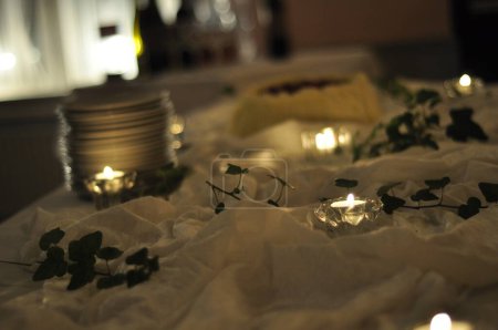 Photo for Arrangement of candles and plants on a table with a white fabric. Tealight candles in clear glass cups in dim lighting and romantic, ambient feeling. Stack of plates and a cake in the background. - Royalty Free Image