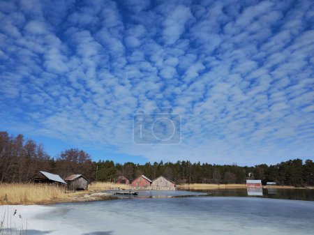 Photo for Frozen lake or sea melting in the spring. Blue sky with unusually shaped soft clouds. Houses, buildings, maybe summer cottages on the shore. Finnish scenic archipelago when winter turns to spring. - Royalty Free Image