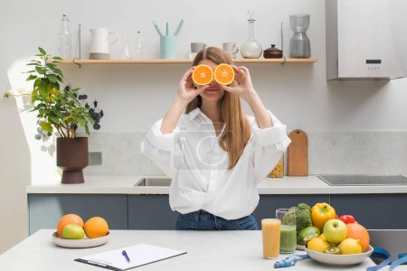 Téléchargez les photos : Une jolie fille sourit et ferme les yeux avec des moitiés orange dans la cuisine. Le concept de bonne nutrition, alimentation, végétarisme. - en image libre de droit