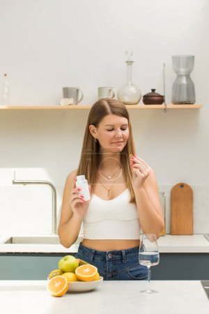 Attractive girl holding vitamin D or omega3 capsule and white jar mockup in kitchen. The concept of food active additives.