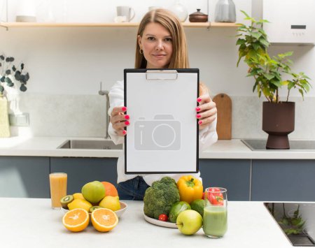 Téléchargez les photos : Jolie fille tient une tablette vierge avec une feuille blanche pour votre texte, debout à la table avec des fruits et légumes frais. Le concept d'un plan de repas, végétarisme. - en image libre de droit