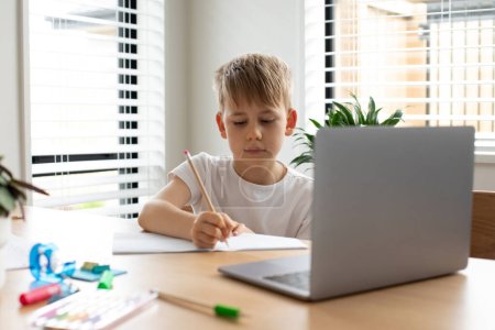 Téléchargez les photos : Un garçon mignon étudie dans une école en ligne. Le garçon est assis devant un ordinateur portable et prend des notes dans un carnet. Le concept d'éducation. - en image libre de droit