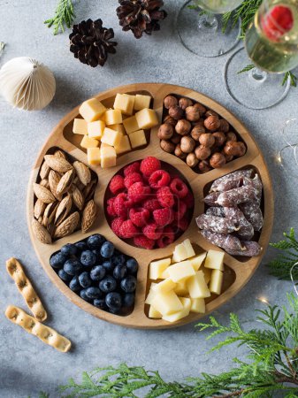 Wooden plate with various snacks on the New Years table. Christmas menu, festive snacks. Assorted cheeses, sausages, nuts in a menazhnitsa.