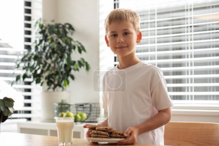 Boy holding a plate of homemade chocolate chip cookies, with a glass of milk on the table. Homemade