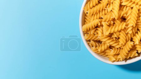 Uncooked fusilli pasta spirals in a white bowl on a blue background, perfect for culinary and food concepts. Copy space