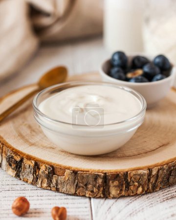 Rich creamy yogurt in a glass bowl on a wooden round, accompanied by fresh blueberries and a golden spoon