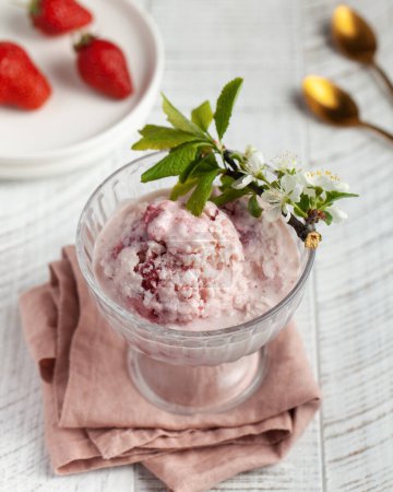 Delicate homemade strawberry ice cream served in an elegant glass bowl, garnished with fresh strawberry slices and a sprig of blossoming cherry. Accompanied by a rustic background with strawberries