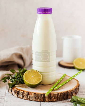 Yogur refrescante con sabor a lima en una botella, presentado en una rebanada de madera con lima fresca y pajitas verdes.