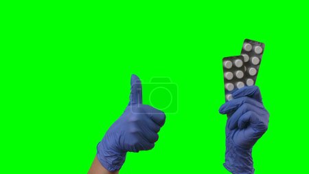 Foto de Las manos de mujer con guantes azules de látex muestran ampollas de pastillas y pulgares hacia arriba. Brazos médicos con guantes médicos protectores que recomiendan medicamentos, vitaminas o antibióticos. Primer plano en la pantalla verde - Imagen libre de derechos