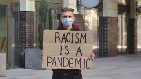 Foto de Hombre joven con máscara médica se para con un póster de cartón RACISMO ES UN PANDÉMICO en un lugar público al aire libre. Un activista político protestando en una protesta. No hay racismo sino tolerancia y corrección política - Imagen libre de derechos