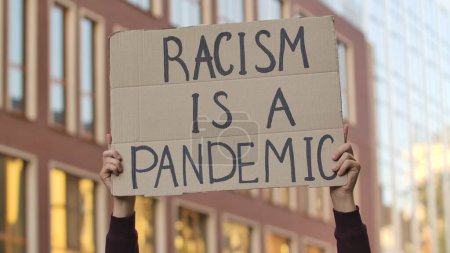 Photo for RACISM IS A PANDEMIC on cardboard poster in hands of male protester activist. Stop Racism concept, No Racism. Rallies against racism and police brutality. Peaceful life of blacks matters. City street - Royalty Free Image
