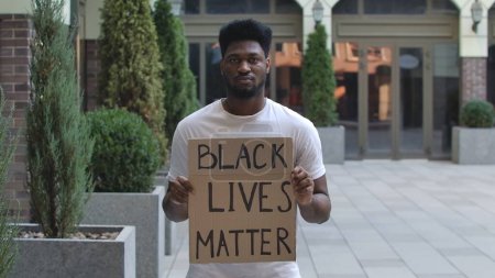 Foto de Joven hombre afroamericano está de pie con un cartel de cartón RACISMO ES UN PANDÉMICO en lugar público al aire libre. Un movimiento antirracista para protestar contra la injusticia y la brutalidad policial. Manifestación callejera - Imagen libre de derechos