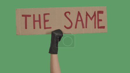 Photo for Hands of an unknown person in black gloves raise cardboard poster THE SAME. Stop Racism concept, No Racism. Rallies against racism and police brutality. Peaceful life of blacks matters. Isolated green - Royalty Free Image