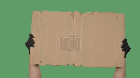 Foto de Las manos en guantes negros levantan un cartel en blanco hecho de una caja de cartón. Espacio vacío para tu texto, logo o anuncio. Concepto de diseño de banner. Aislado una pantalla verde, croma clave - Imagen libre de derechos