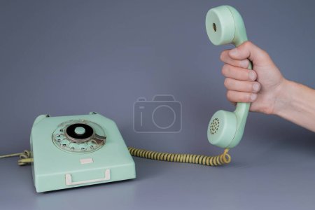 Téléchargez les photos : Main masculine tenant un vieux récepteur de téléphone en plastique bleu près du téléphone rotatif sur fond gris. Fermez le combiné à distance de l'appareil de téléphone domestique rétro dans les mains de l'homme. Concept de communication - en image libre de droit