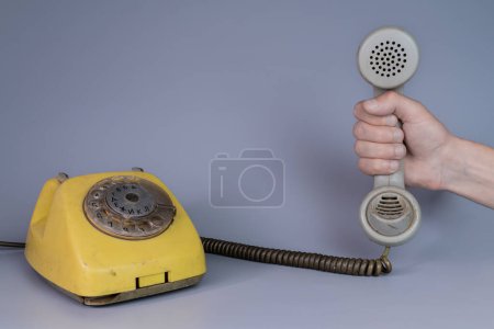 Téléchargez les photos : Main masculine tenant un vieux récepteur de téléphone en plastique près du téléphone rotatif jaune sur fond gris. Fermez le combiné à distance de l'appareil de téléphone domestique rétro dans les mains de l'homme. Notion de - en image libre de droit