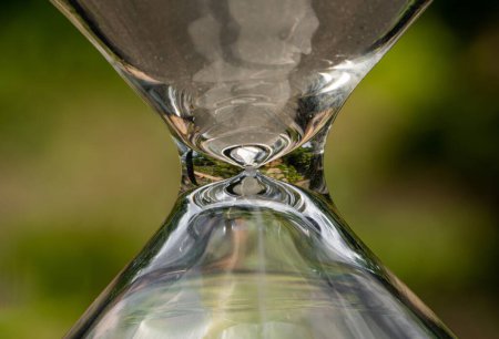 Photo for Macro shot of a glass hourglass case against a blurred green nature background. Vessel of transparent glassy sandglass with reflective backdrop of green garden or country house. Timers counting down - Royalty Free Image