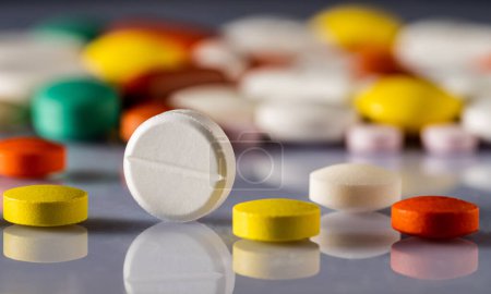 Different colored pills and tablets scattered on a glossy reflective surface. Close up of white, yellow, brown and green medicines. Antibiotics, vitamins, painkillers. Background of medical