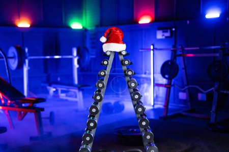 Foto de Bastidor con conjunto de mancuernas de metal en forma de árbol de Navidad y sombrero de Santa Claus rojo. Equipamiento deportivo en gimnasio con luz azul. Sombrillas, pesas y bancos deportivos para crossfit, musculación, fitness - Imagen libre de derechos