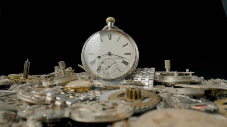Photo for Old silver pocketwatch on a pile of gears, gearwheels, cogwheels and a metal watch bracelet on black background. Disassembled parts clockwork of antique clocks in workshop, repair. Broken clock - Royalty Free Image