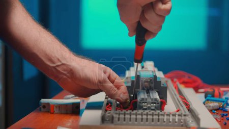 Foto de Unrecognizable man with a screwdriver in his hands twists the red wire in an automatic electrical switch in an electrical workshop. An electrician is repairing a switchboard with switches. Close up - Imagen libre de derechos