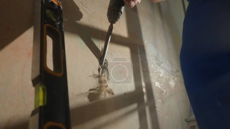 Foto de Professional worker makes a strobe in the wall using a puncher to install an electrical cable. Close up of man in blue construction overalls cutting concrete wall with plaster. Concept of repair - Imagen libre de derechos