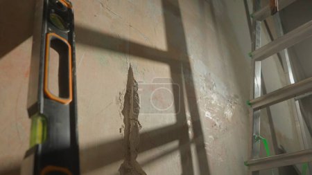 Photo for Strobe in the wall for laying wiring or pipes. A damaged hole in a concrete wall, a water level and a metal ladder close up. The shadow of the ladder falls on the wall. Concept of repair, construction - Royalty Free Image