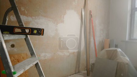 Téléchargez les photos : Black level lying on metal ladder against brown wall. The interior of the room inside in the process of renovation or construction work. Cardboard boxes are covered with polyethylene. Measuring - en image libre de droit