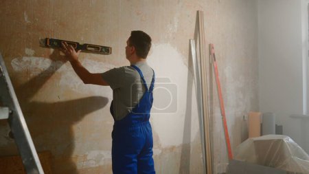 Foto de A man in blue construction overalls is measuring a wall with a water level. Room with ladder, boxes and pile of polyethylene. Concept of repair, construction - Imagen libre de derechos