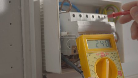 Foto de Work under voltage, electrician is checking serviceability of equipment, measuring voltage by yellow multimeter. Close up of a mans hands testing white switch box check voltage switchboard. Search for - Imagen libre de derechos