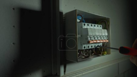 Photo for Electrician assembles electrical panel in dark by light of flashlight. Close up of a man hands tightening a screw with a screwdriver in a switchboard high voltage electrical switches. Switch box with - Royalty Free Image