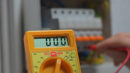 Foto de Yellow multimeter for measuring voltage close up. Work under voltage, electrician is checking serviceability of equipment. Mans hands testing white switch box check voltage switchboard. Search for - Imagen libre de derechos