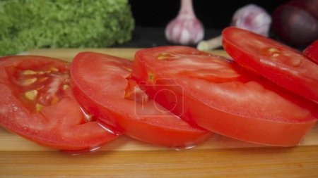 Photo for Juicy sliced rings of ripe red tomato with water drops on wooden board. Wet tomato slices with juicy pulp, green salad, garlic and onion on black background. Fresh salad ingredients. Macro shot - Royalty Free Image