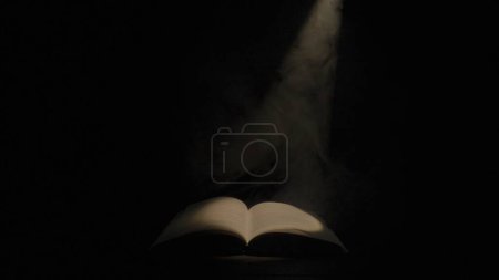 Photo for An open book illuminated by a beam of light and smoke on a black isolated studio background. An old textbook with a spot of light on the pages in the darkness of the library. Mysterious open book - Royalty Free Image