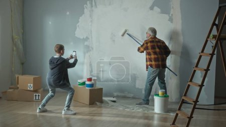 Photo for Grandson takes a photo with mobile phone or filming a video of grandpa painting a wall white with a paint roller. Elderly man doing repairs in an apartment and posing for photo in memory. Concept of - Royalty Free Image