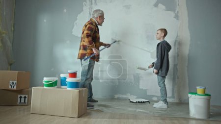 Téléchargez les photos : Grand-père enseigne à son petit-fils comment peindre un mur avec un rouleau de peinture et donne des conseils précieux. Un homme âgé et un adolescent font des réparations dans leur appartement sur fond de carton - en image libre de droit