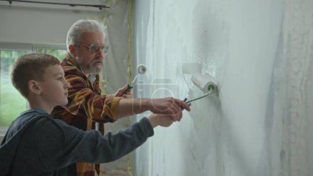 Photo for Side view of grandfather and grandson painting wall with white paint using paint rollers. An elderly man teaching a young guy and giving advice. The concept of family, teamwork, apartment renovation - Royalty Free Image