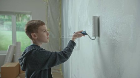 Téléchargez les photos : Jeune garçon en sweat à capuche bleu peint un mur blanc avec rouleau de peinture. Adolescent examinant un mur et exécutant diligemment un rouleau le long du mur. Concept de réparation dans l'appartement, décoration, apprentissage - en image libre de droit