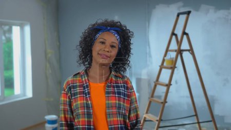 Téléchargez les photos : Young African American woman is looking at the camera and smiling. Portrait of black female with curly hair in checkered shirt is posing against the backdrop of an apartment in process of renovation - en image libre de droit