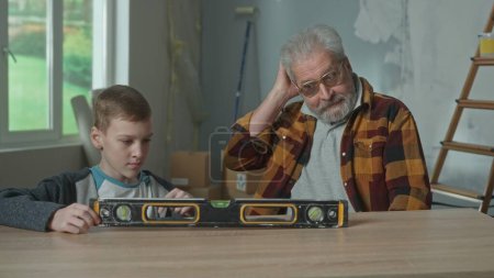 Téléchargez les photos : Grandpa and grandson are studying the principle of operation of the water level, which lies on the table. A teenager examines the instrument with interest, and an elderly man scratches his head in - en image libre de droit