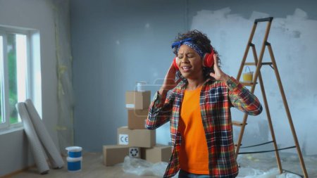 Photo for African American woman in big wireless red headphones is enjoying music. Black female in checkered shirt sings with closed eyes in apartment against the backdrop of the ladder, cardboard boxes and - Royalty Free Image